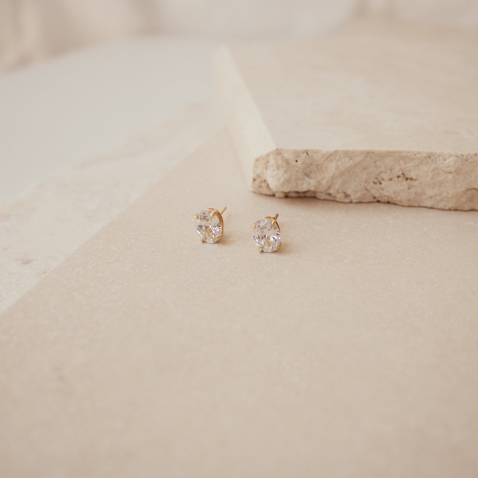 The Oval Diamond Studs are classic gold earrings featuring clear gemstones, displayed on a beige stone surface.