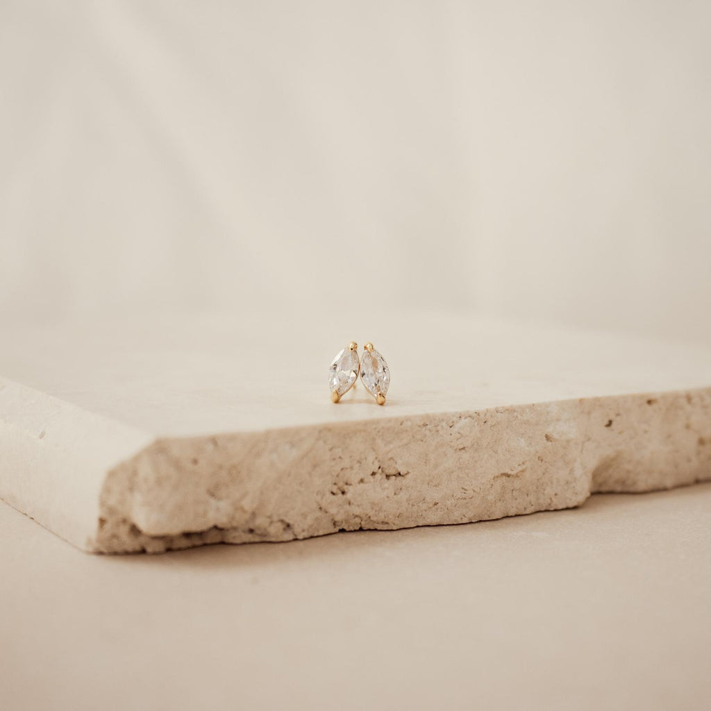The Classic Marquise Diamond Studs shine with elegance in gold settings on a textured stone slab, presenting a modern update to timeless jewelry.