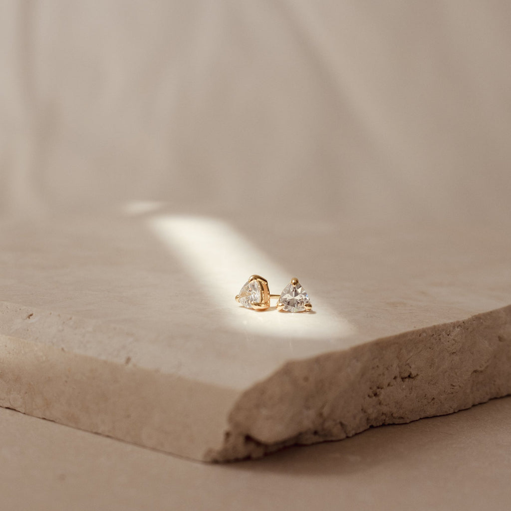 The Triangle Diamond Studs, featuring a gold triangle design with sparkling diamonds, are displayed on a textured stone slab under soft natural light for a timeless, classic jewelry look.