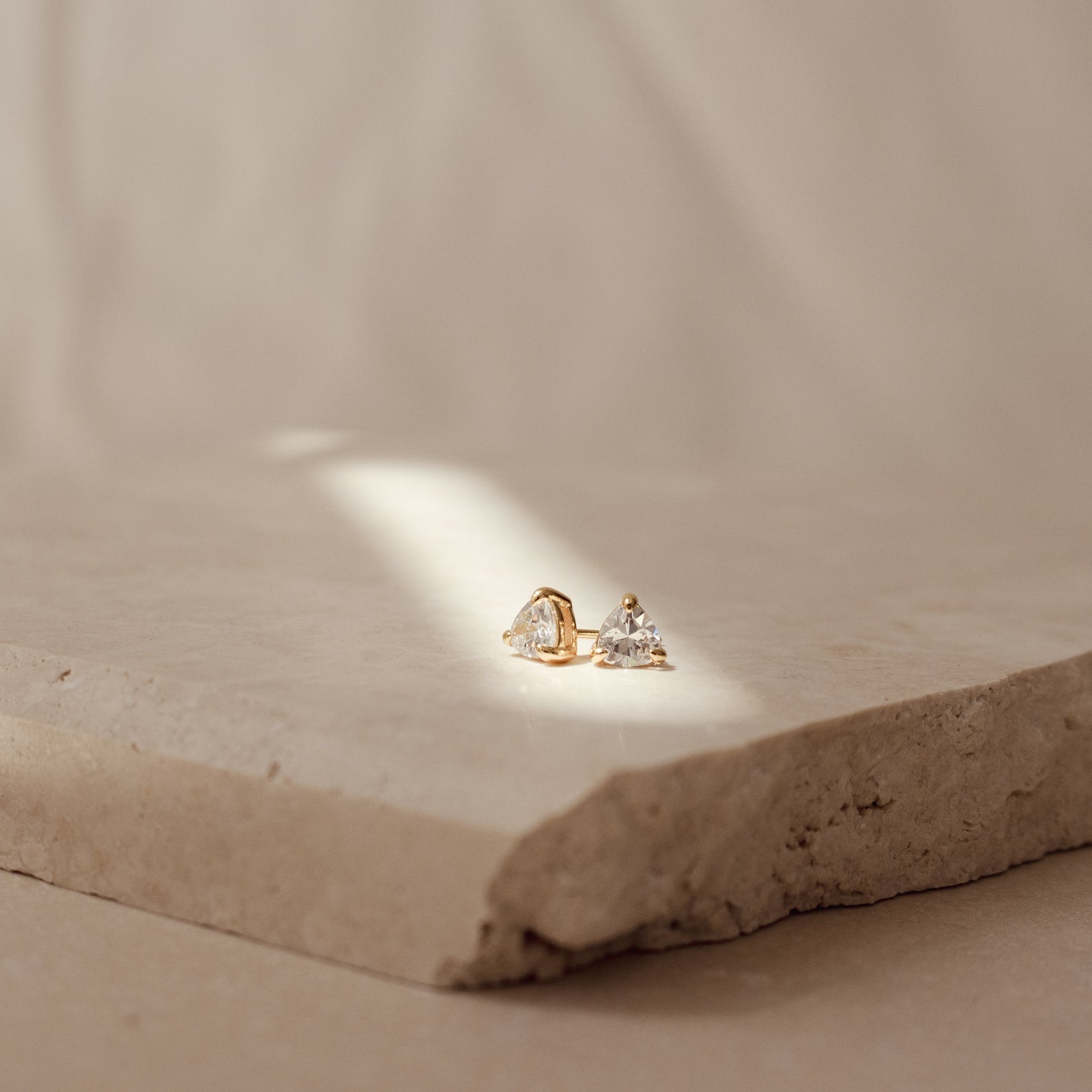 The Triangle Diamond Studs, featuring a gold triangle design with sparkling diamonds, are displayed on a textured stone slab under soft natural light for a timeless, classic jewelry look.