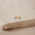 Triangle Diamond Studs, featuring clear triangle-shaped stones set in gold, are a stunning example of classic jewelry, displayed on a textured beige stone surface.