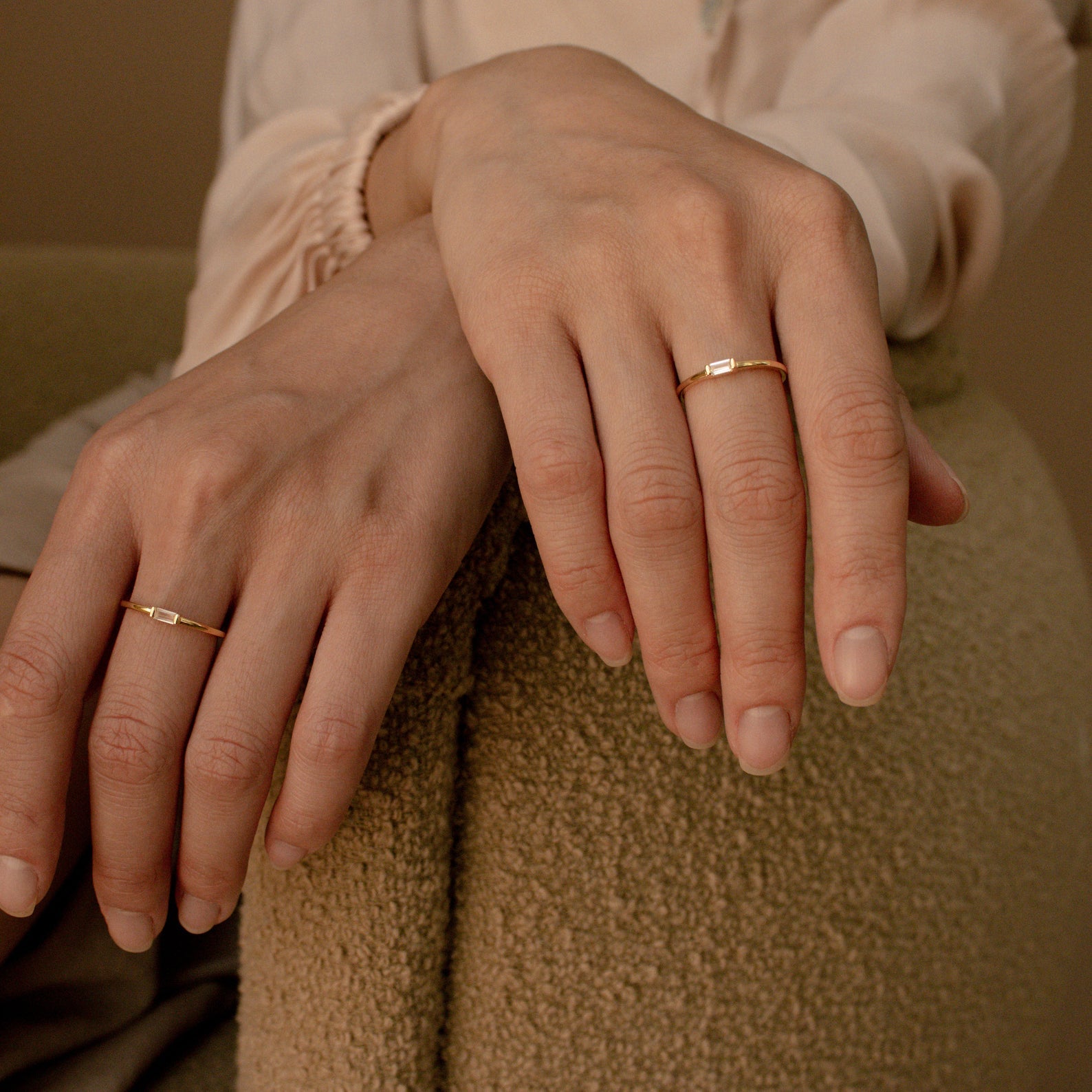 Close-up of two hands with neatly manicured nails, each wearing a Baguette Diamond Ring, resting on a textured chair.