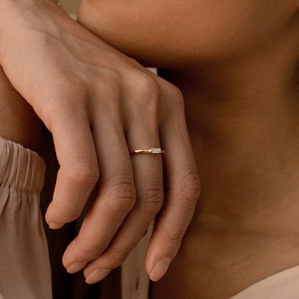A close-up shows a hand wearing the Baguette Diamond Ring, featuring a delicate gold band and an east-west set baguette diamond, resting near the face.