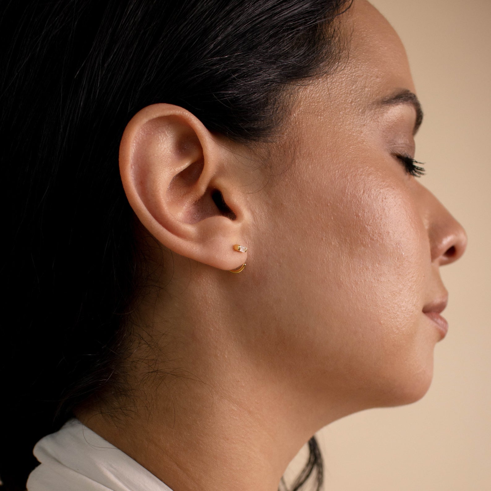 A woman with closed eyes, shown in profile against a neutral background, wears Shayla Baguette Huggies—elegant diamond hoop earrings.