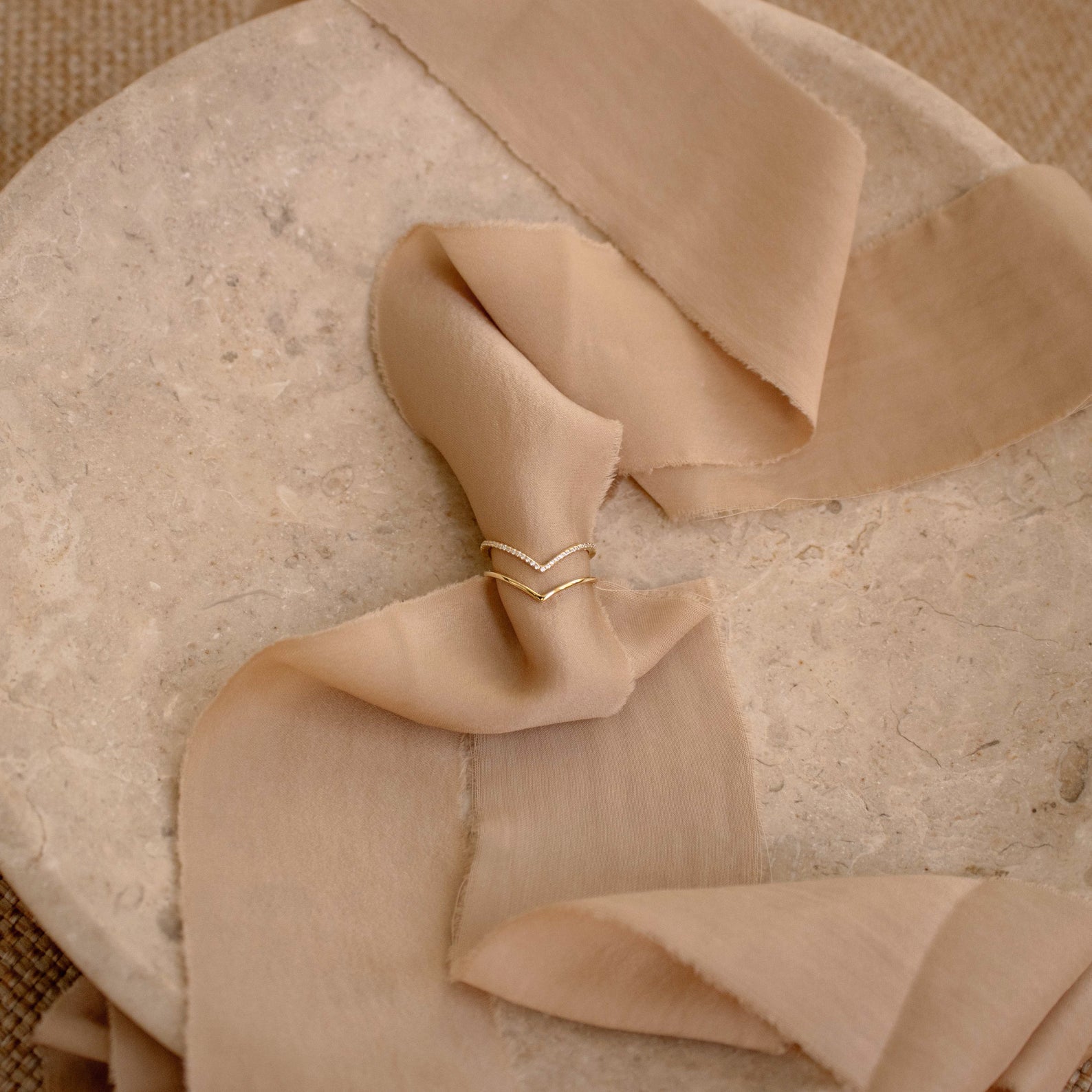 The Curve Stacking Set features gold chevron curved bands elegantly wrapped around a soft beige ribbon, showcased on a textured stone surface.