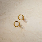 The Maggie Pearl Drop Hoops feature modern oval pearls hanging from sleek hoops, shown on a light beige, textured surface—ideal for minimal jewelry lovers.