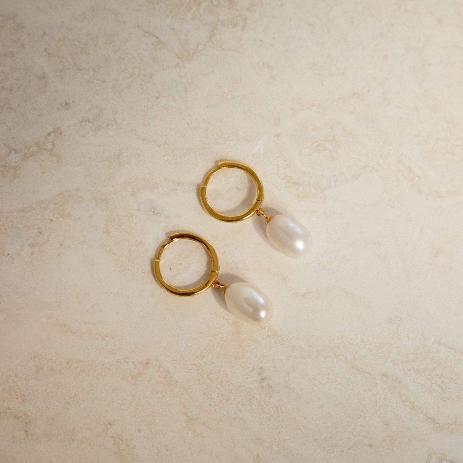 The Maggie Pearl Drop Hoops feature modern oval pearls hanging from sleek hoops, shown on a light beige, textured surface—ideal for minimal jewelry lovers.