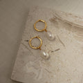 Maggie Pearl Drop Hoops—gold hoops with dangling white pearls—are shown on a light textured stone surface, making them the perfect minimal jewelry piece.