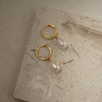 Maggie Pearl Drop Hoops—gold hoops with dangling white pearls—are shown on a light textured stone surface, making them the perfect minimal jewelry piece.