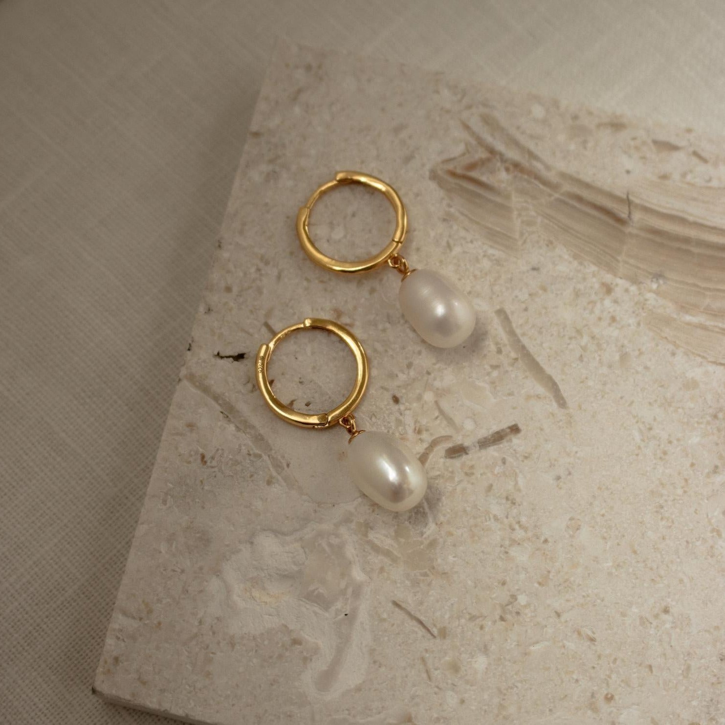 Maggie Pearl Drop Hoops—gold hoops with dangling white pearls—are shown on a light textured stone surface, making them the perfect minimal jewelry piece.