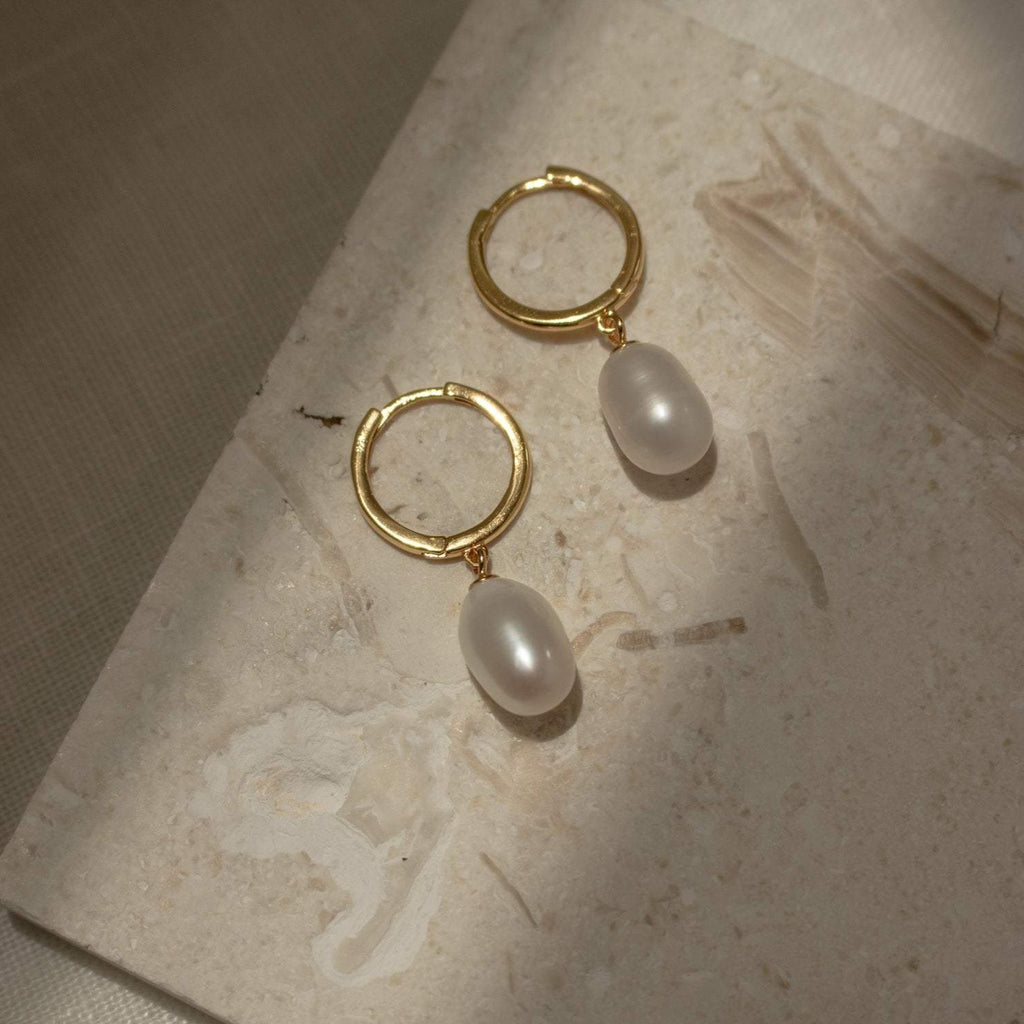 Maggie Pearl Drop Hoops—modern hoop earrings featuring dangling oval pearls, showcased on a textured beige stone, ideal for minimal jewelry lovers who appreciate timeless style.