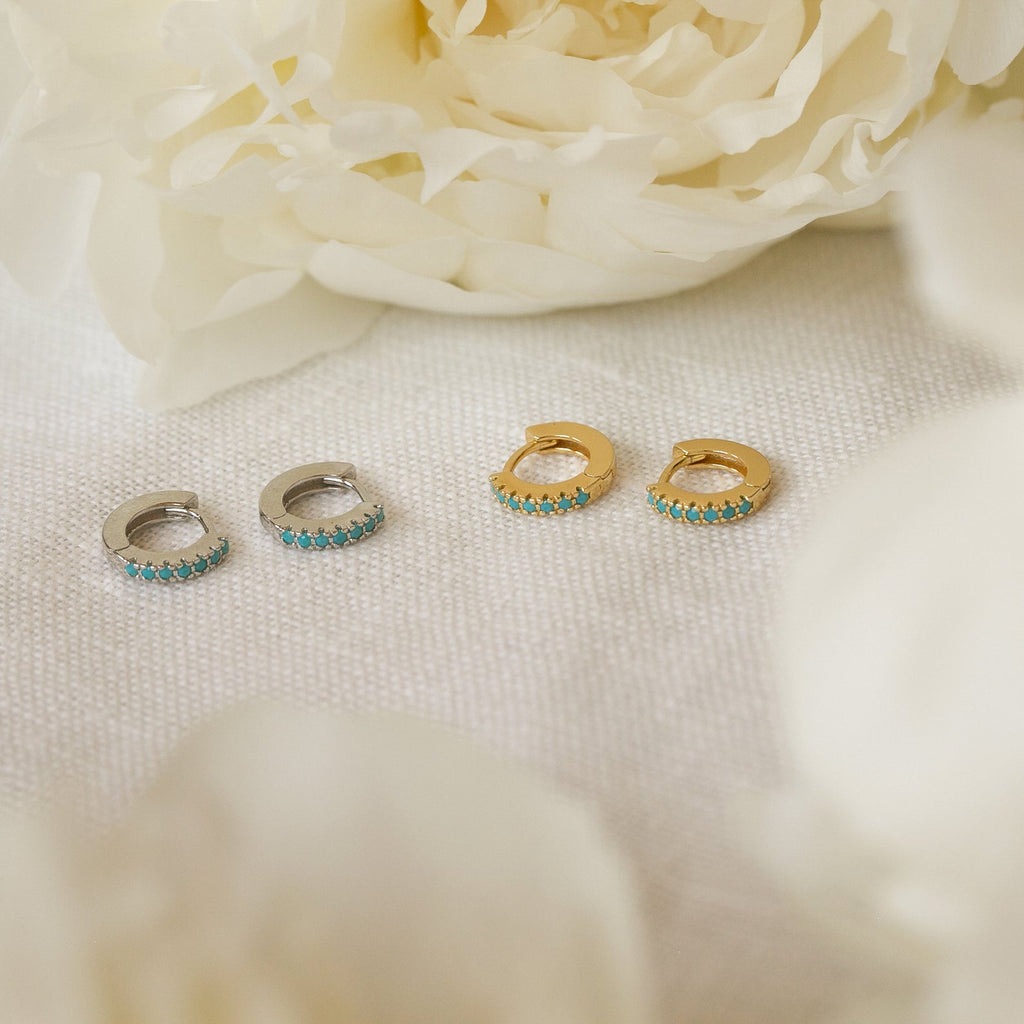 The Kendra Huggies, available in silver and gold pairs, are displayed on white fabric with flowers—blending eclectic charm and ideal for layering to create your own unique style.