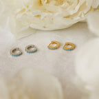 The Kendra Huggies, available in silver and gold pairs, are displayed on white fabric with flowers—blending eclectic charm and ideal for layering to create your own unique style.