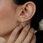 Close-up of a person’s ear adorned with Kendra Huggies—a small gold hoop earring featuring green stones and eclectic charm; hand near the ear.