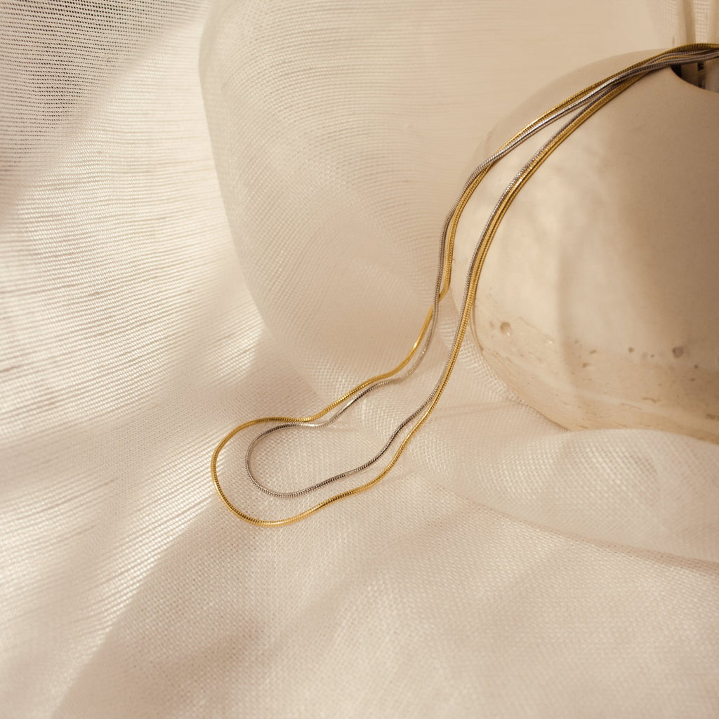 A slightly wider view of the same dual-tone snake chains (gold and white gold) resting on textured ivory fabric with a neutral stone.