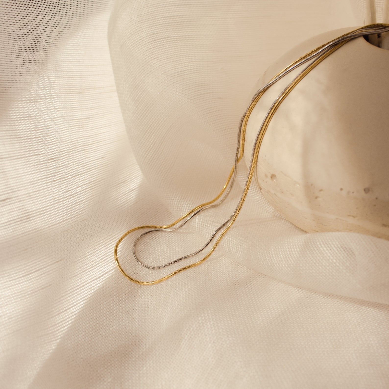 A slightly wider view of the same dual-tone snake chains (gold and white gold) resting on textured ivory fabric with a neutral stone.