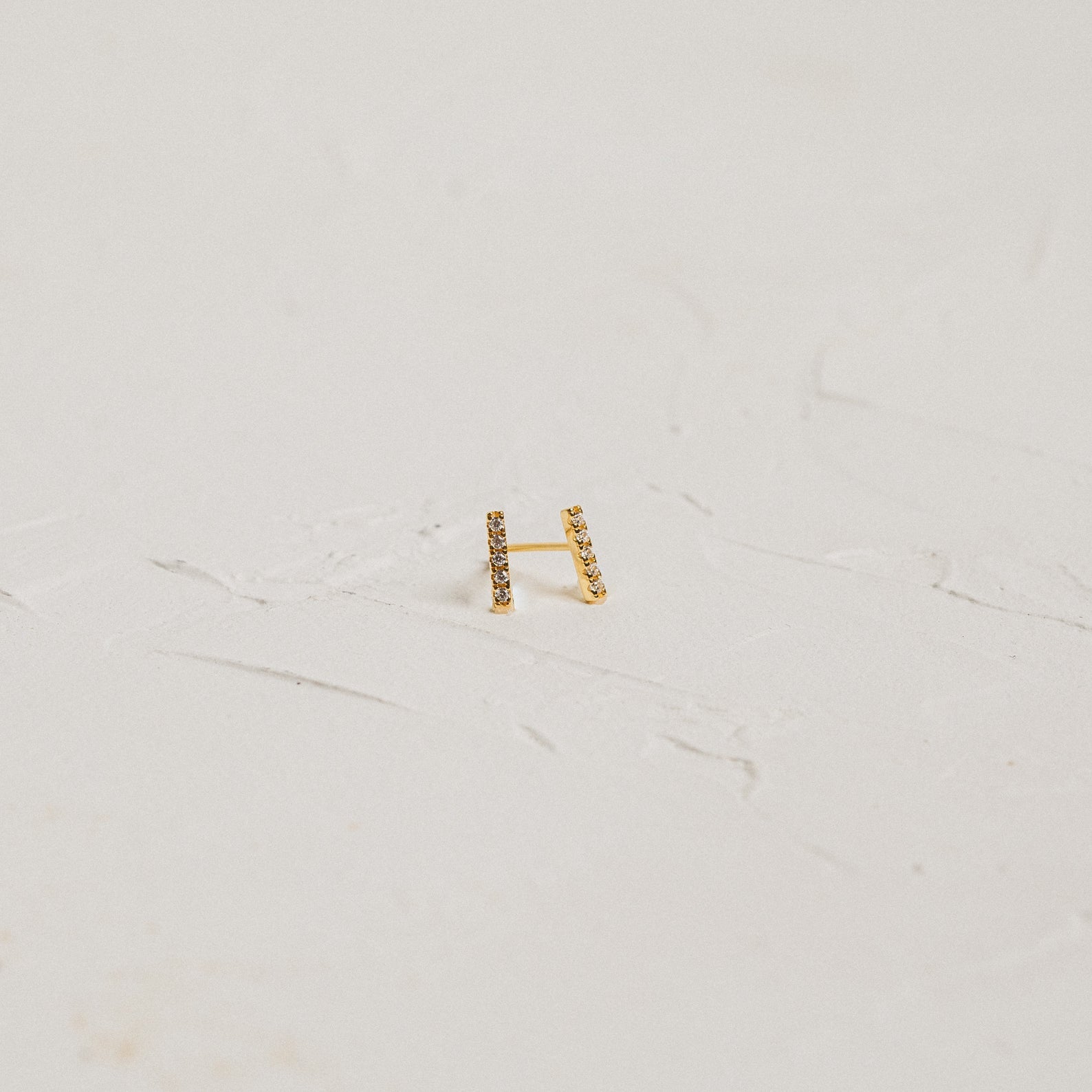 Minimal gold Pave Bar Studs with embedded stones rest elegantly on a white textured surface.