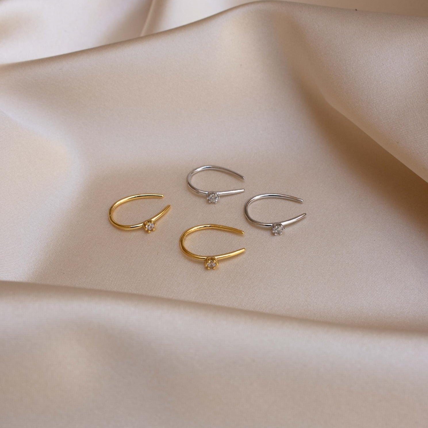 Four Rylee Diamond Wire Earrings—two gold and two silver, each adorned with small gems—rest on smooth beige fabric.