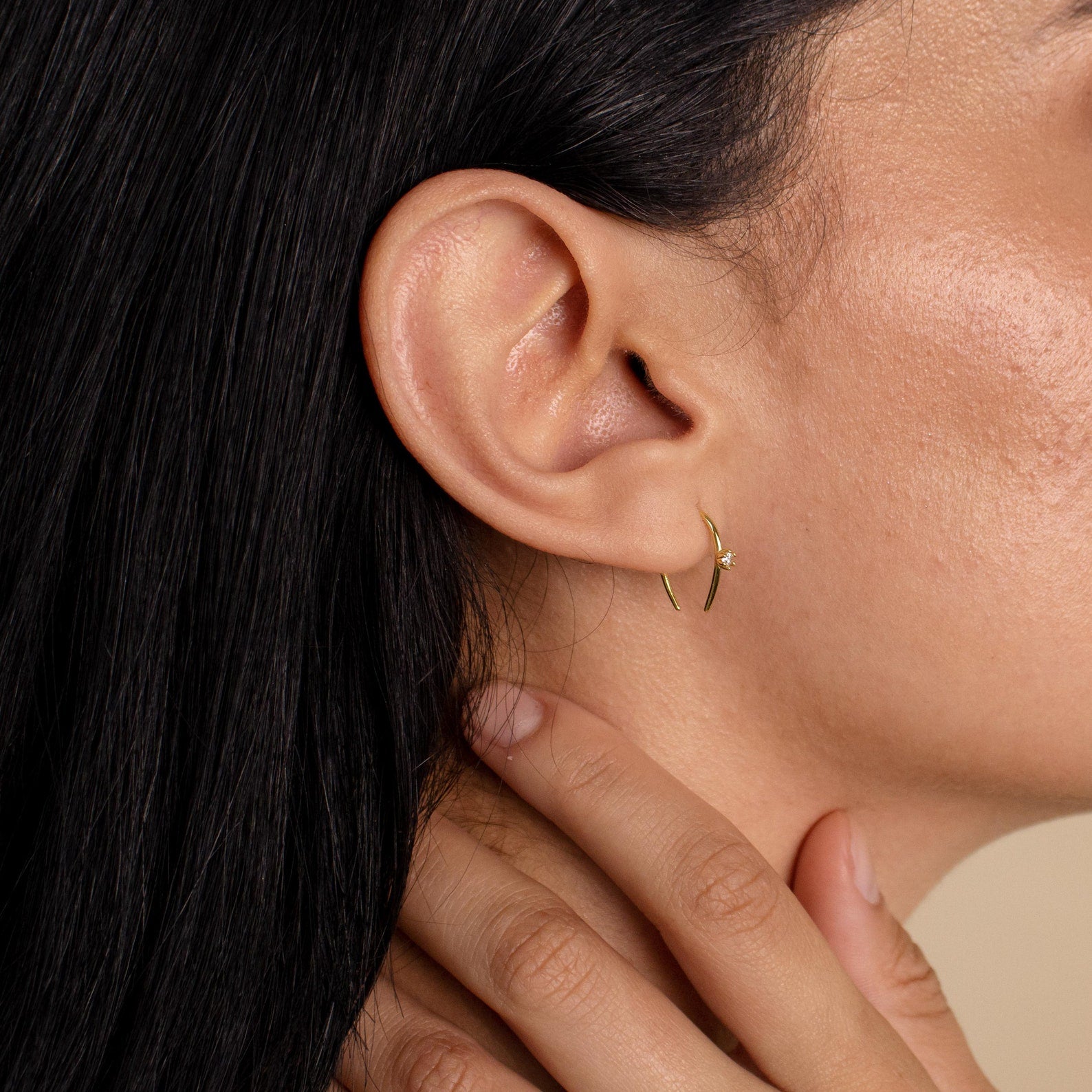 Close-up of a woman’s ear wearing the Rylee Diamond Wire Earrings, a delicate gold open hoop design, as her hand rests gently on her neck.