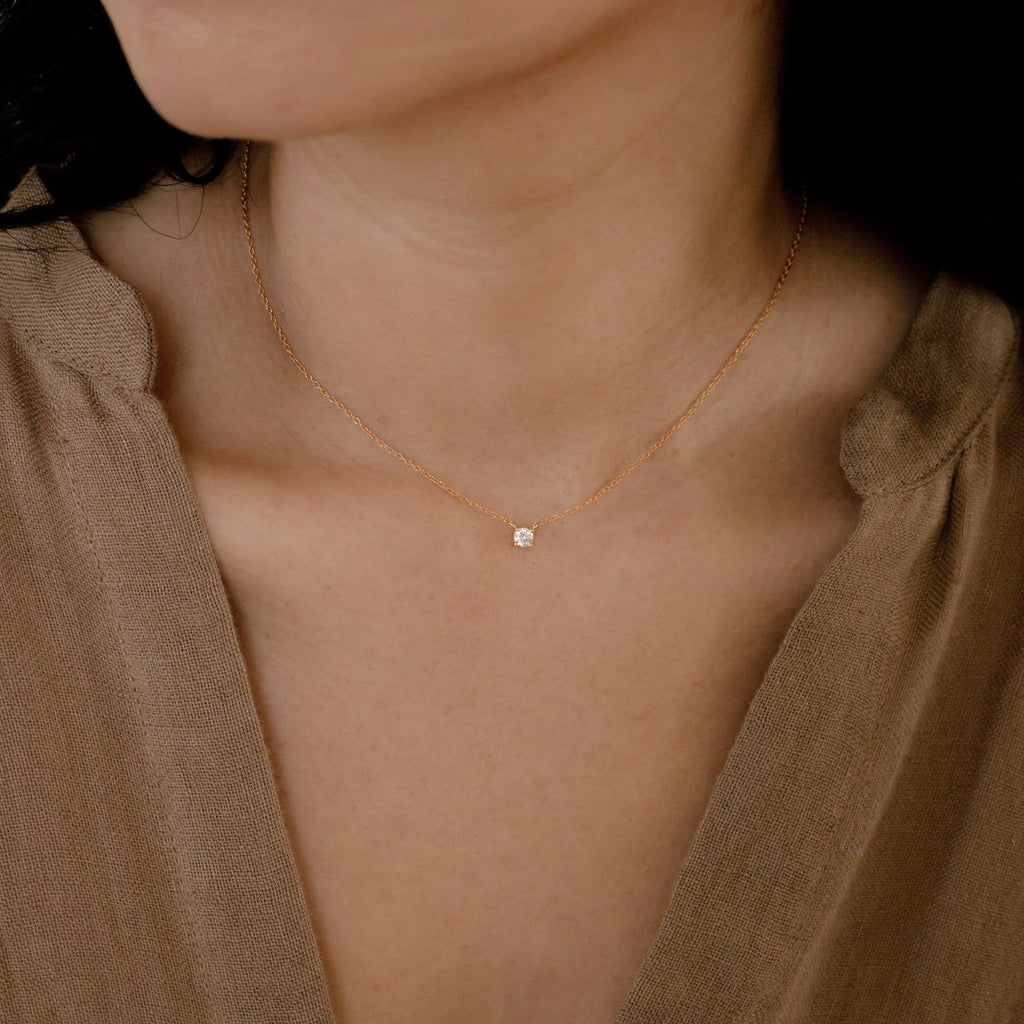 A woman models the Dainty Diamond Necklace, featuring a small diamond charm pendant set in 925 sterling silver, against her bare neck and a beige v-neck top.