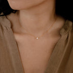A woman models the Dainty Diamond Necklace, featuring a small diamond charm pendant set in 925 sterling silver, against her bare neck and a beige v-neck top.