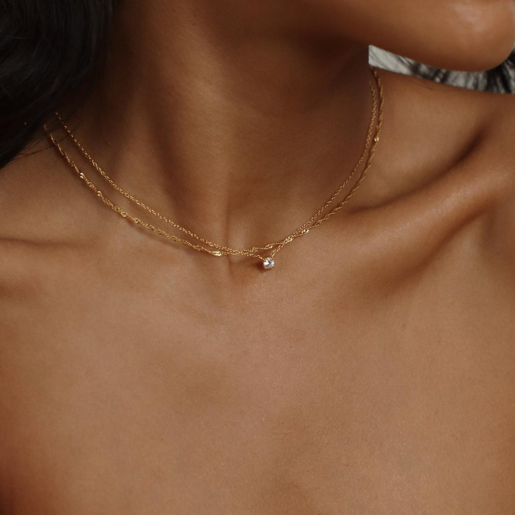 A close-up of a womans neck and collarbone showcases the Dainty Diamond Necklace—a delicate gold chain with a small diamond charm—while a blurred background highlights the jewelry.