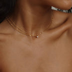 A close-up of a womans neck and collarbone showcases the Dainty Diamond Necklace—a delicate gold chain with a small diamond charm—while a blurred background highlights the jewelry.