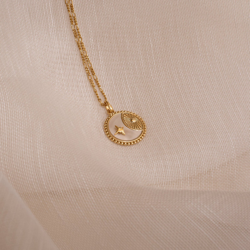 Gold celestial coin necklace with a round pendant featuring a crescent moon, star, and sunburst design, resting on a sheer beige fabric background.