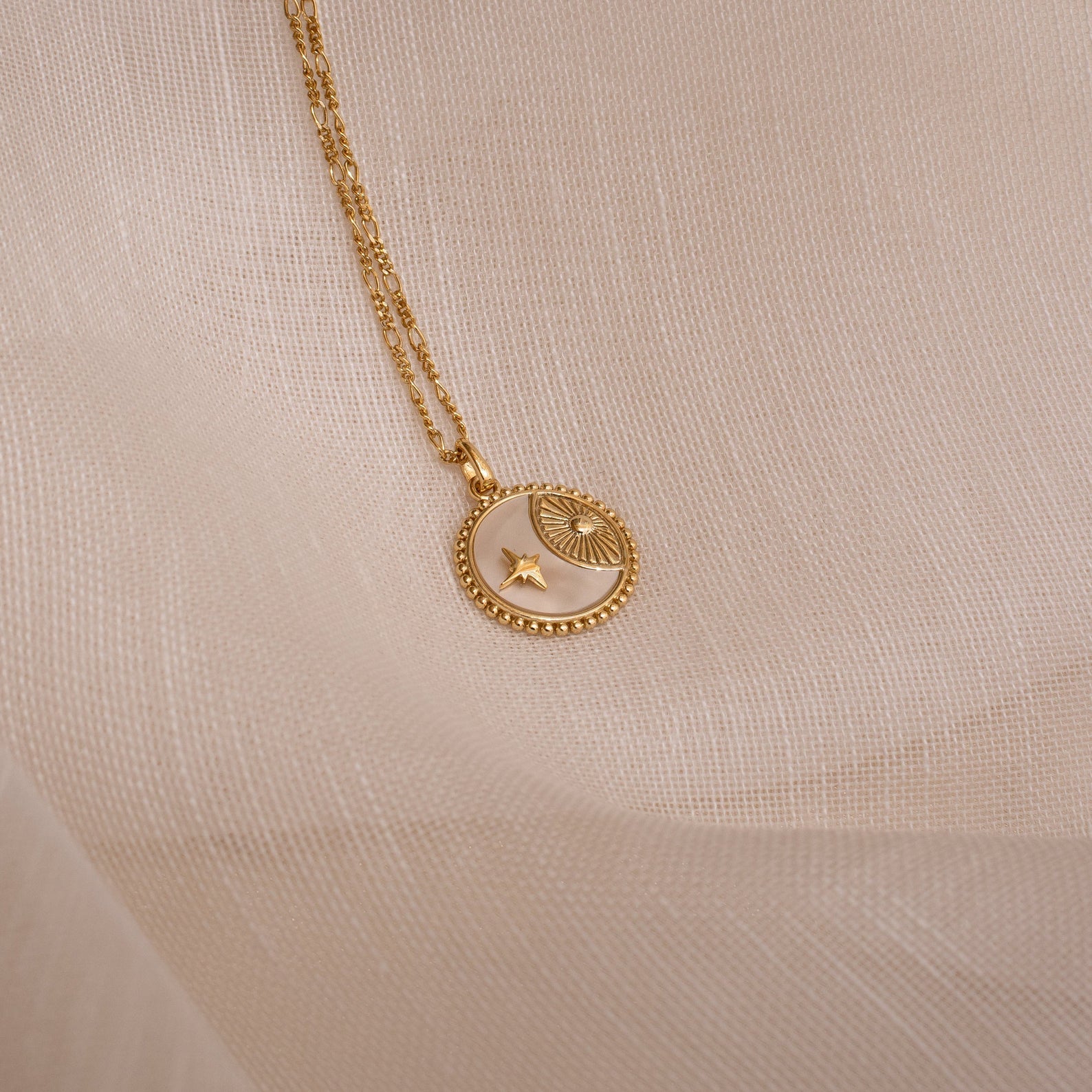 Gold celestial coin necklace with a round pendant featuring a crescent moon, star, and sunburst design, resting on a sheer beige fabric background.