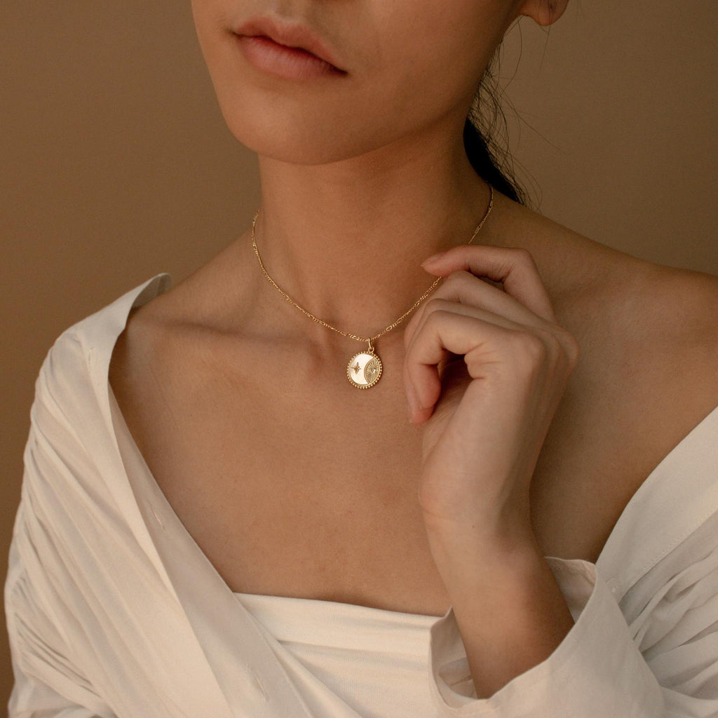 A woman in an off-shoulder white top gently touches the minimal Gabi Coin Necklace, showcasing its simple gold design.