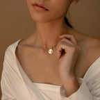 A woman in an off-shoulder white top gently touches the minimal Gabi Coin Necklace, showcasing its simple gold design.