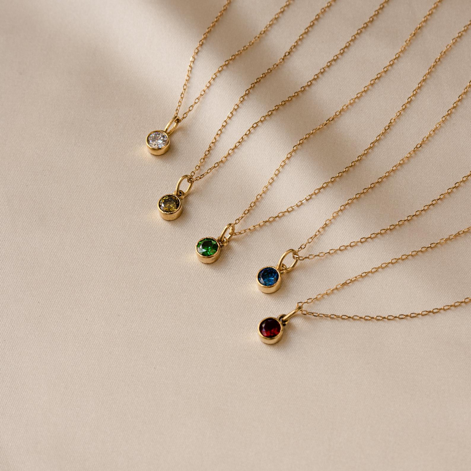 The Custom Birthstone Necklace features five delicate gold chains with round gemstone pendants in assorted colors on a beige backdrop—an ideal personalized jewelry gift or meaningful birthstone keepsake.