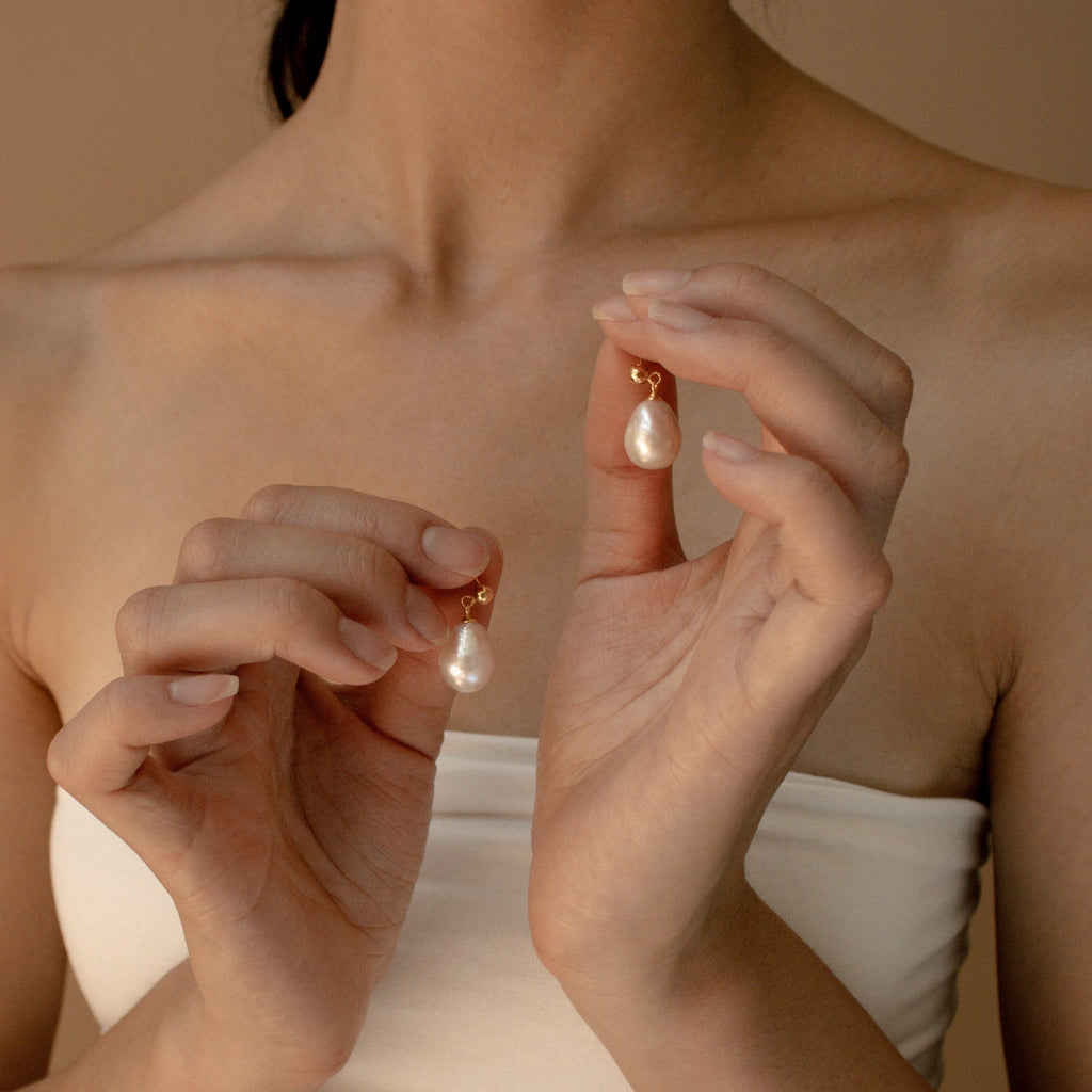 A woman in a white top holds the Baroque Pearl Earrings, showcasing their large statement design near her collarbone.