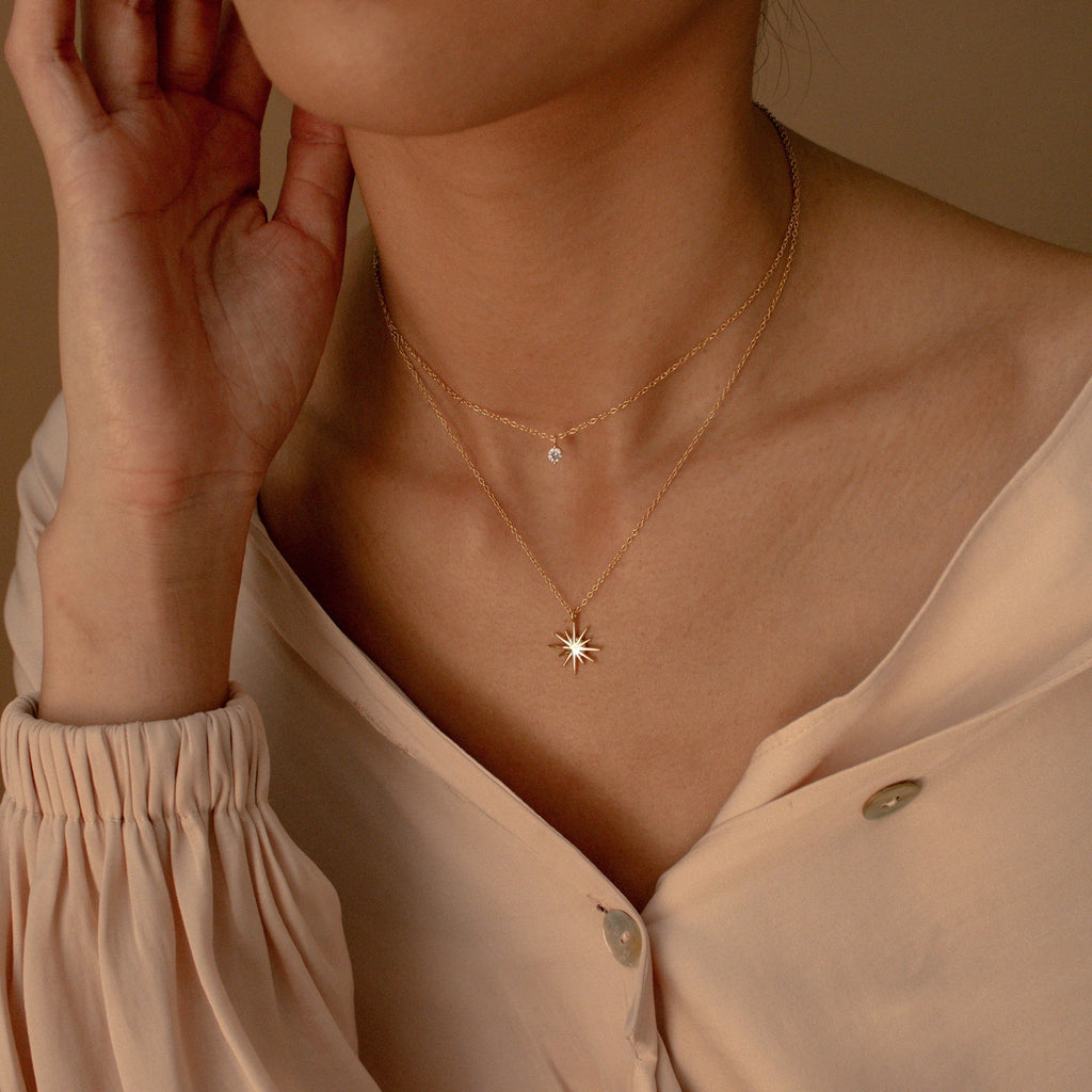 A person wearing a cream blouse with the Laura Starburst Duo Necklace in 18K Gold—featuring one diamond-accented chain and another with a star-shaped pendant.