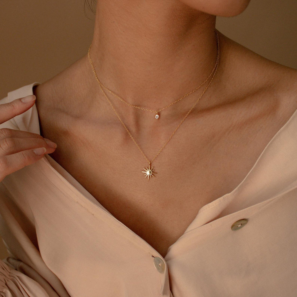 A woman in a light, off-shoulder blouse wears the Laura Starburst Duo Necklace in 18K Gold, featuring one diamond-accented chain and another with a star-shaped pendant.
