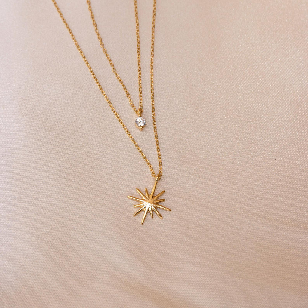 The Laura Starburst Duo Necklace in 18K Gold is displayed on light beige fabric, featuring two gold chains—one with a diamond-shaped pendant and the other with a starburst pendant set with a central gemstone.
