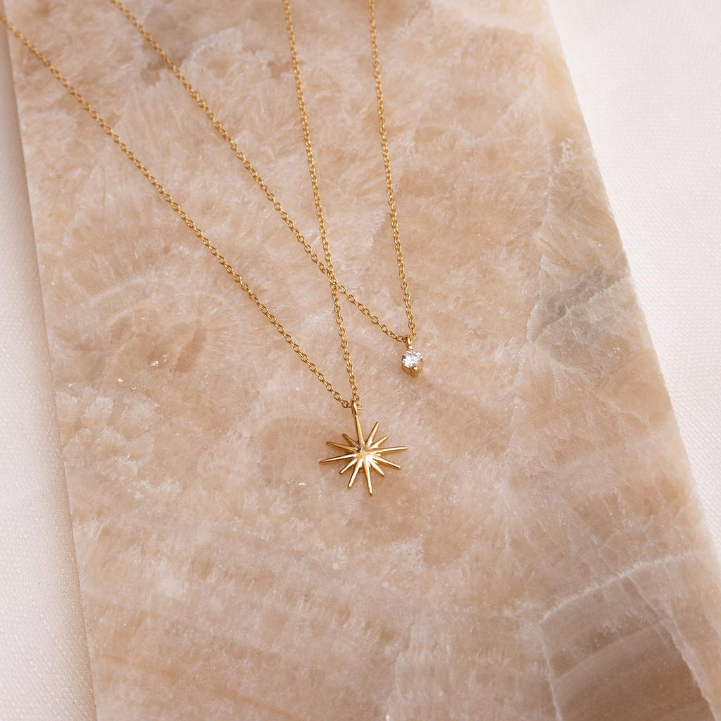 The Laura Starburst Duo Necklace in 18K Gold, featuring two delicate gold chains—a starburst pendant and a diamond accent with a clear gemstone—rests on a beige marble surface.