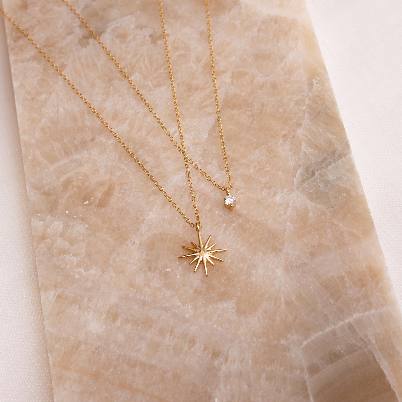 The Laura Starburst Duo Necklace in 18K Gold, featuring two delicate gold chains—a starburst pendant and a diamond accent with a clear gemstone—rests on a beige marble surface.