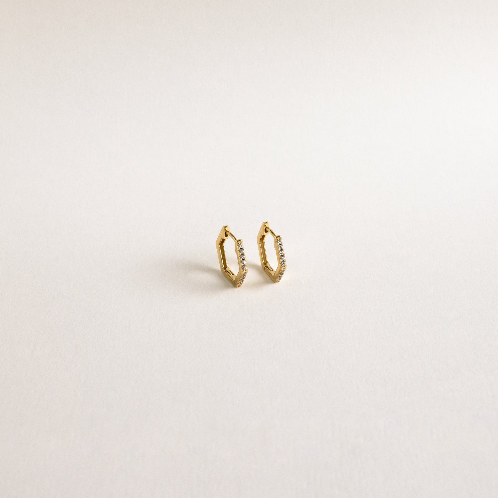 The Parker Pave Hexagon Huggies are small gold earrings with diamond accents, elegantly designed to shine brilliantly against a plain white background.