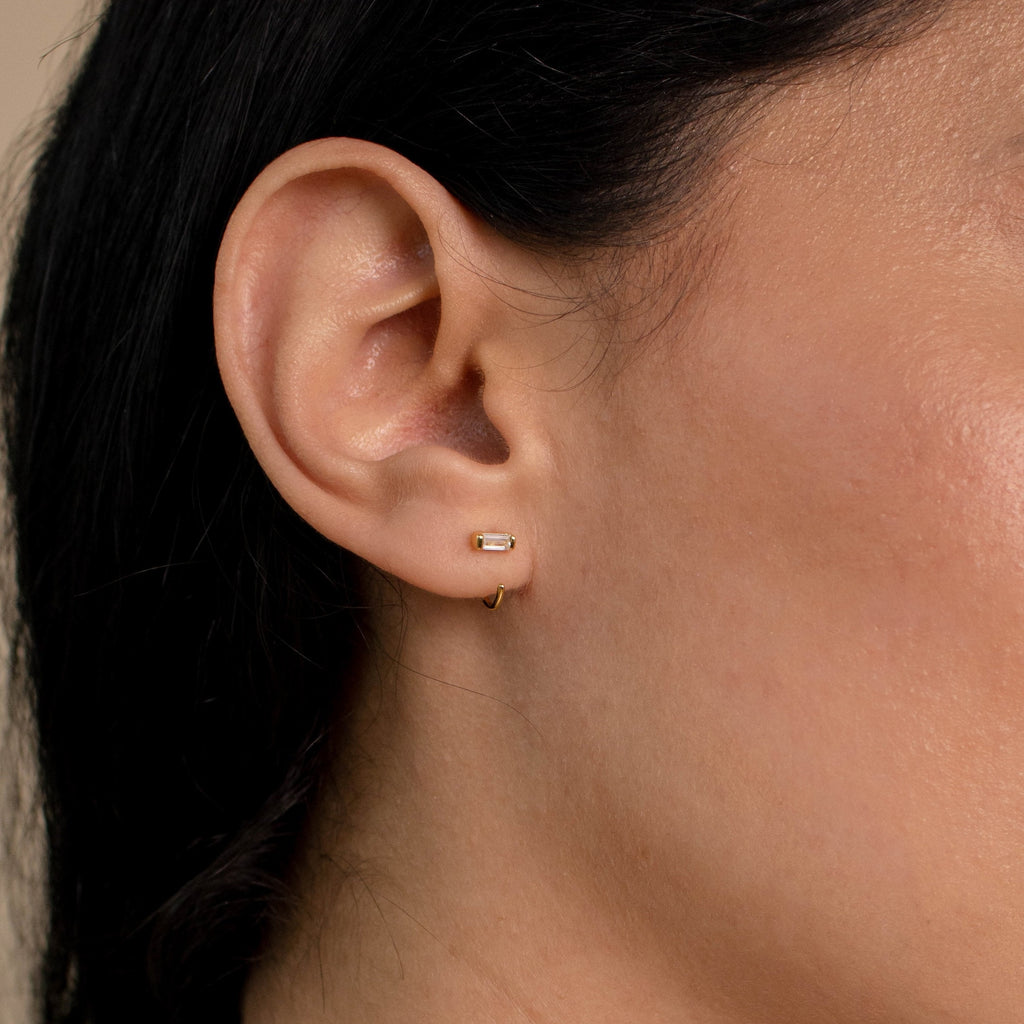 Close-up of a woman's ear adorned with two small gold earrings—a stud and the Shayla Baguette Huggies, featuring a delicate baguette-shaped diamond.