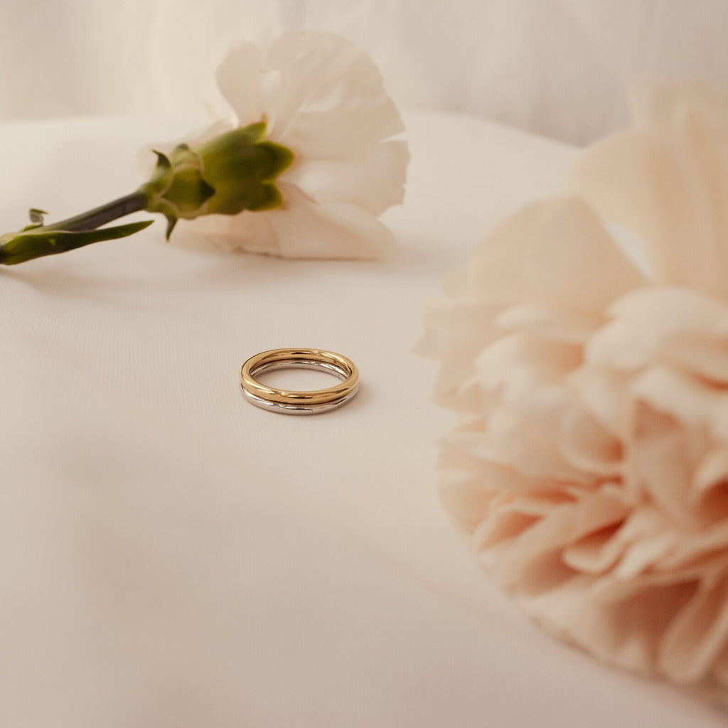 A Minimal Stacking Ring rests on a white cloth, surrounded by softly blurred pale pink carnations.