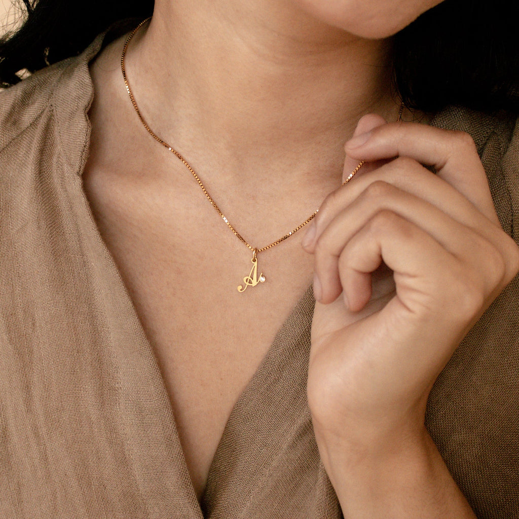 A woman holds the Initial Birthstone Necklace with an A pendant—a personalized gold piece—near her collarbone.