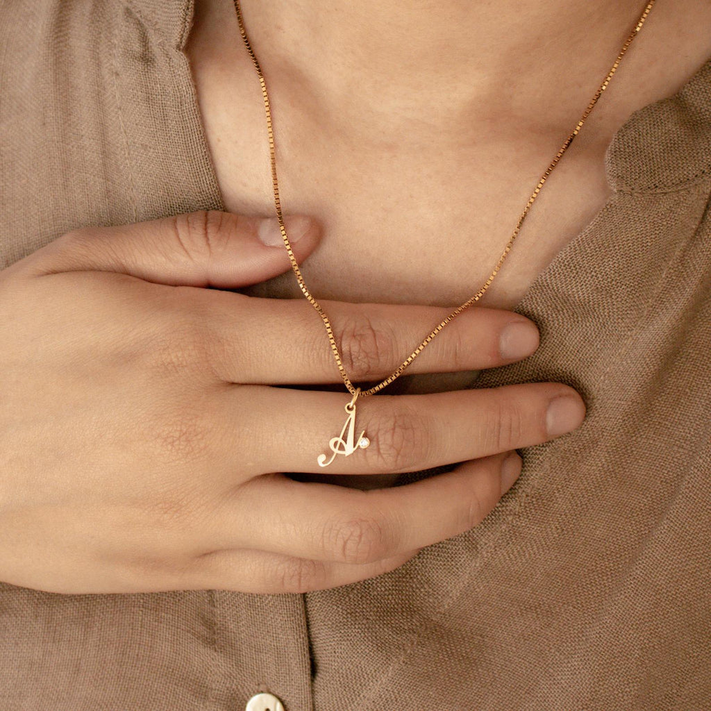 A hand holds the Initial Birthstone Necklace featuring a gold A pendant, styled over a beige shirt—a personalized gift ideal for any occasion.