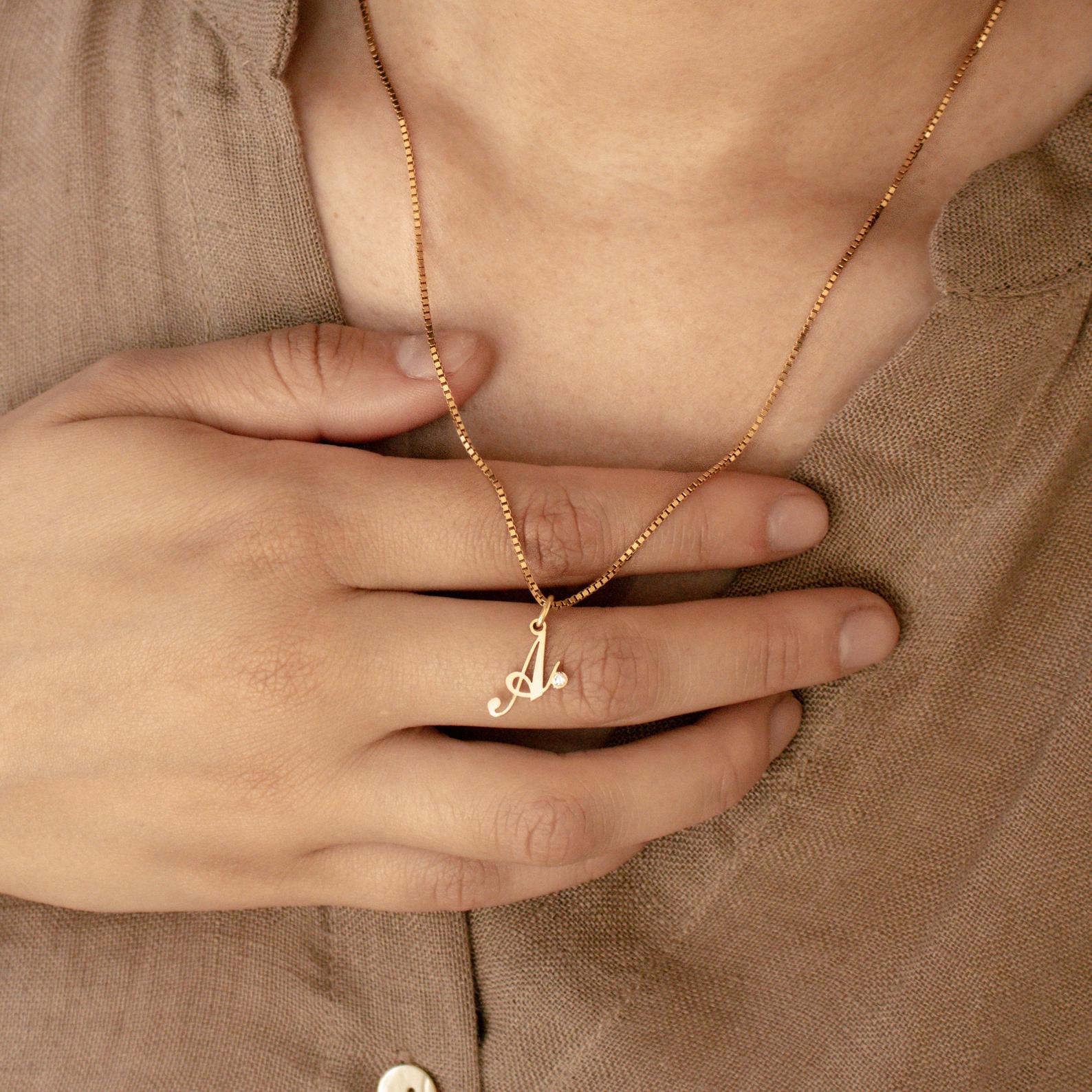 A hand holds the Initial Birthstone Necklace featuring a gold A pendant, styled over a beige shirt—a personalized gift ideal for any occasion.