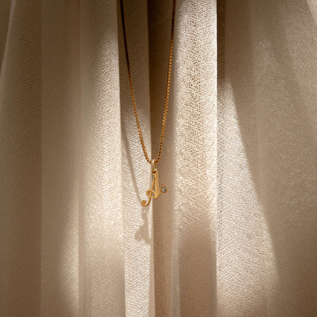 The Initial Birthstone Necklace pendant glows in sunlight against cream-colored curtains—a perfect personalized gift for any occasion.