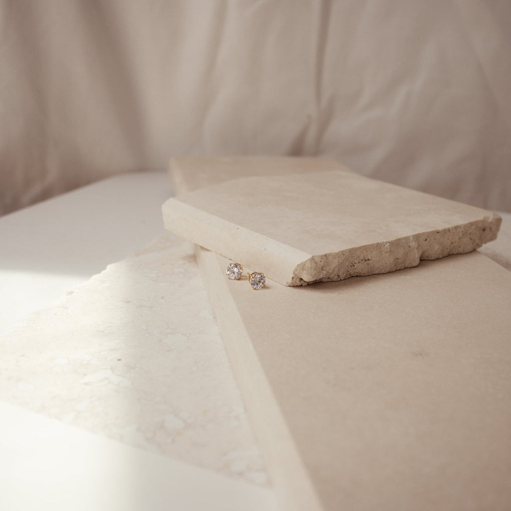 The Round Diamond Studs are displayed on beige stone slabs against a soft, neutral background.
