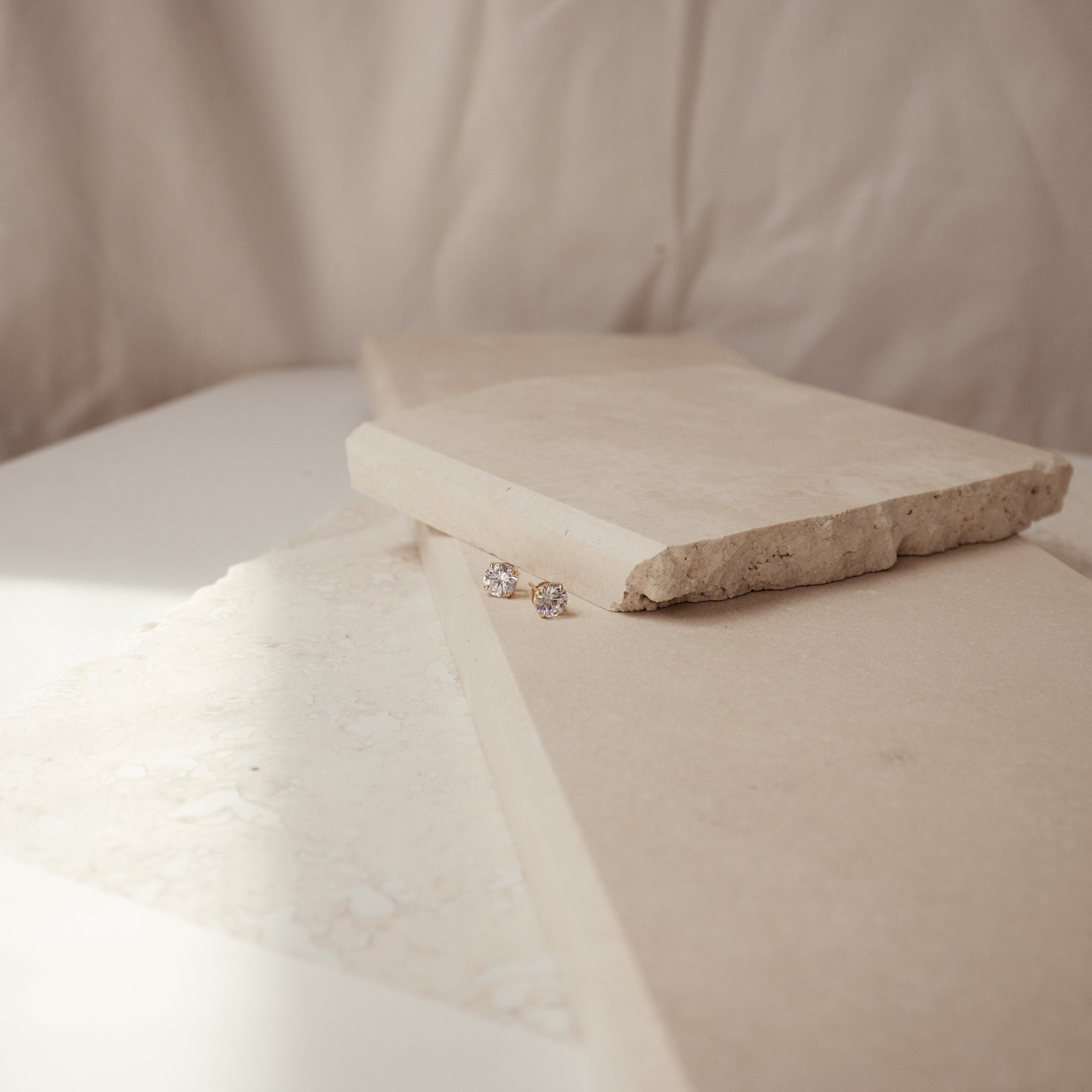 The Round Diamond Studs are displayed on beige stone slabs against a soft, neutral background.
