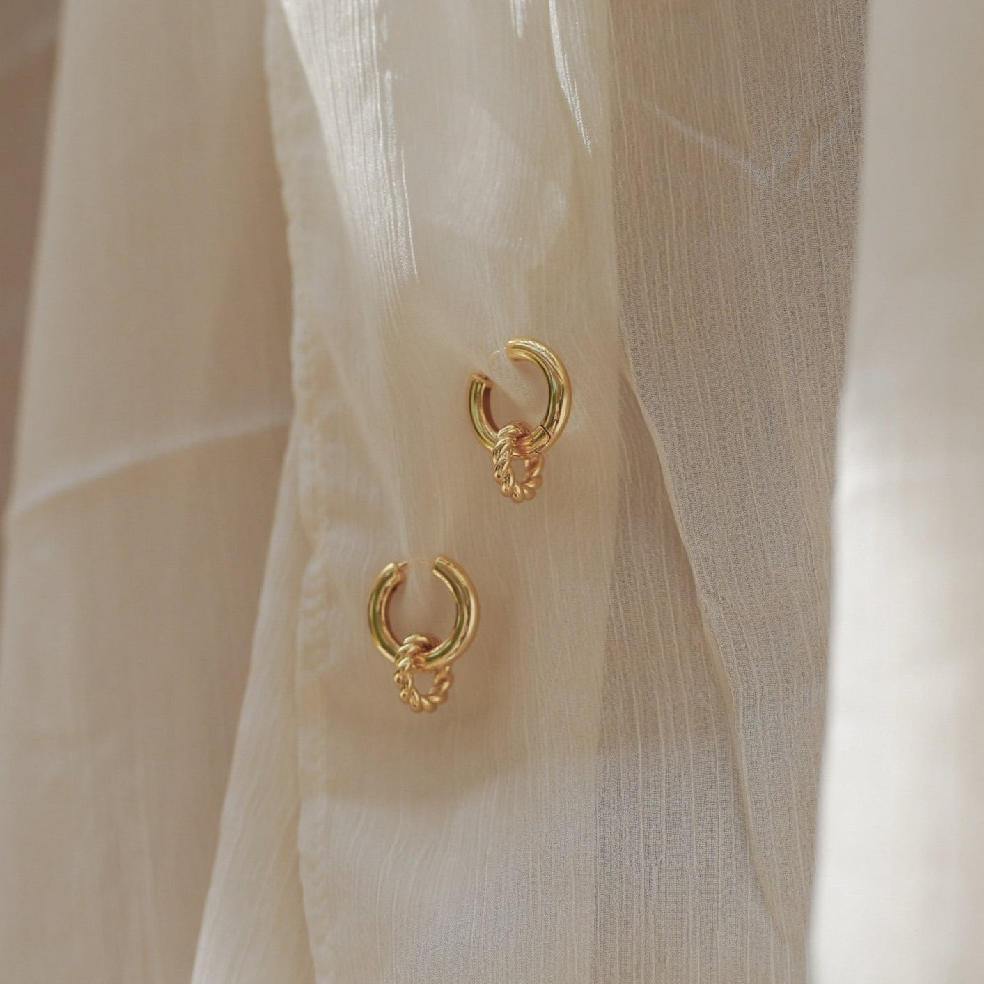 Two Claire Duo Hoops with small charms are displayed on a sheer, cream-colored fabric—ideal for adding chic flair to any outfit.