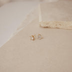 The Oval Diamond Studs are classic gold earrings featuring oval clear gemstones, perfect for lovers of timeless jewelry, shown on a beige stone surface.