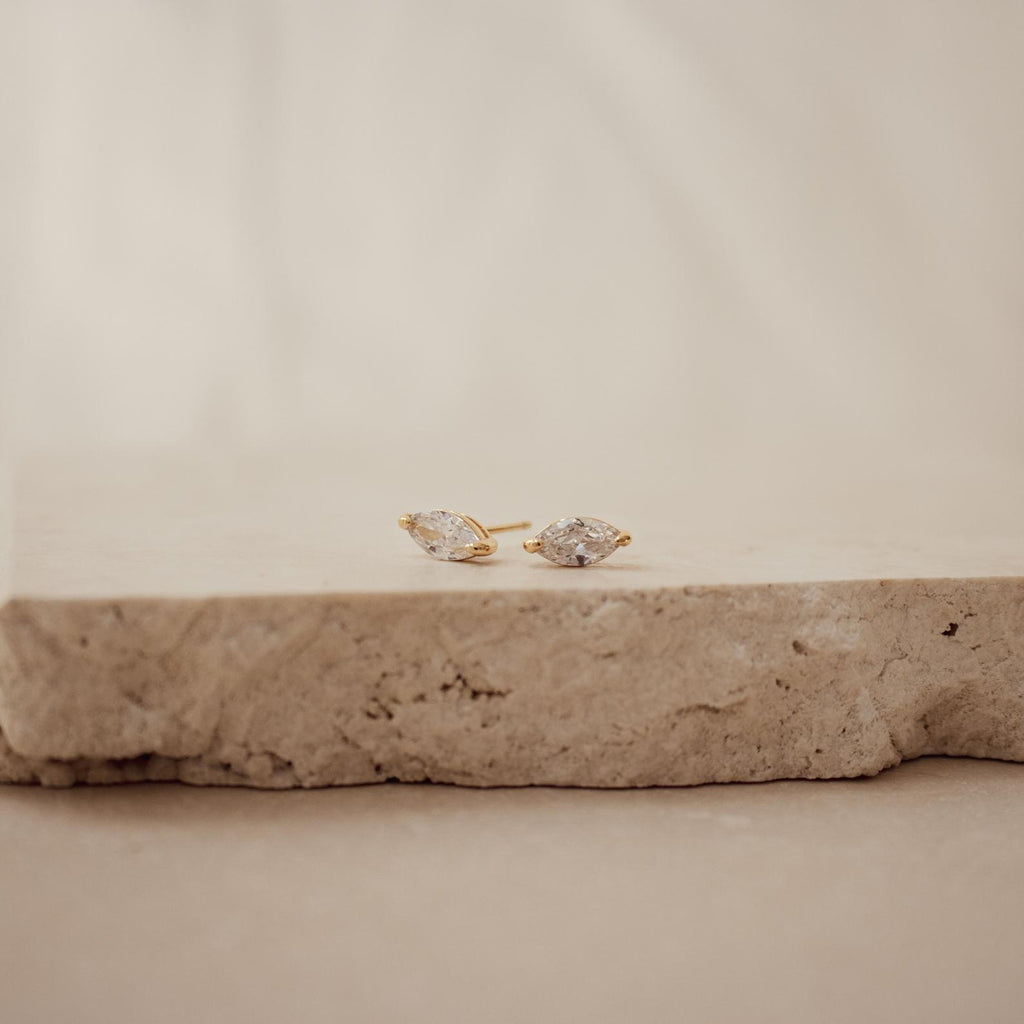 Classic Marquise Diamond Studs, featuring marquise-cut clear stones in a timeless gold setting, are elegantly displayed on a textured beige stone surface.